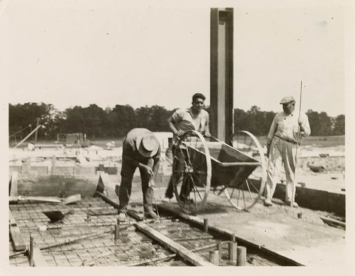 Photograph of construction workers building Jordan Hall. On the left, a worker is bent over adjusting the rebar foundation, in the center, a man pushes a wheelbarrow, and on the right, a man watches the other two workers. 