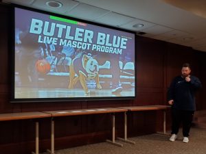 Projected slide showing words "Butler Blue Live Mascot Program" and Evan Krauss standing to the right holding a microphone