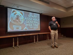 Chief Conley stands with microphone to the right of a projected slide reading "Staff Assembly General Meeting"